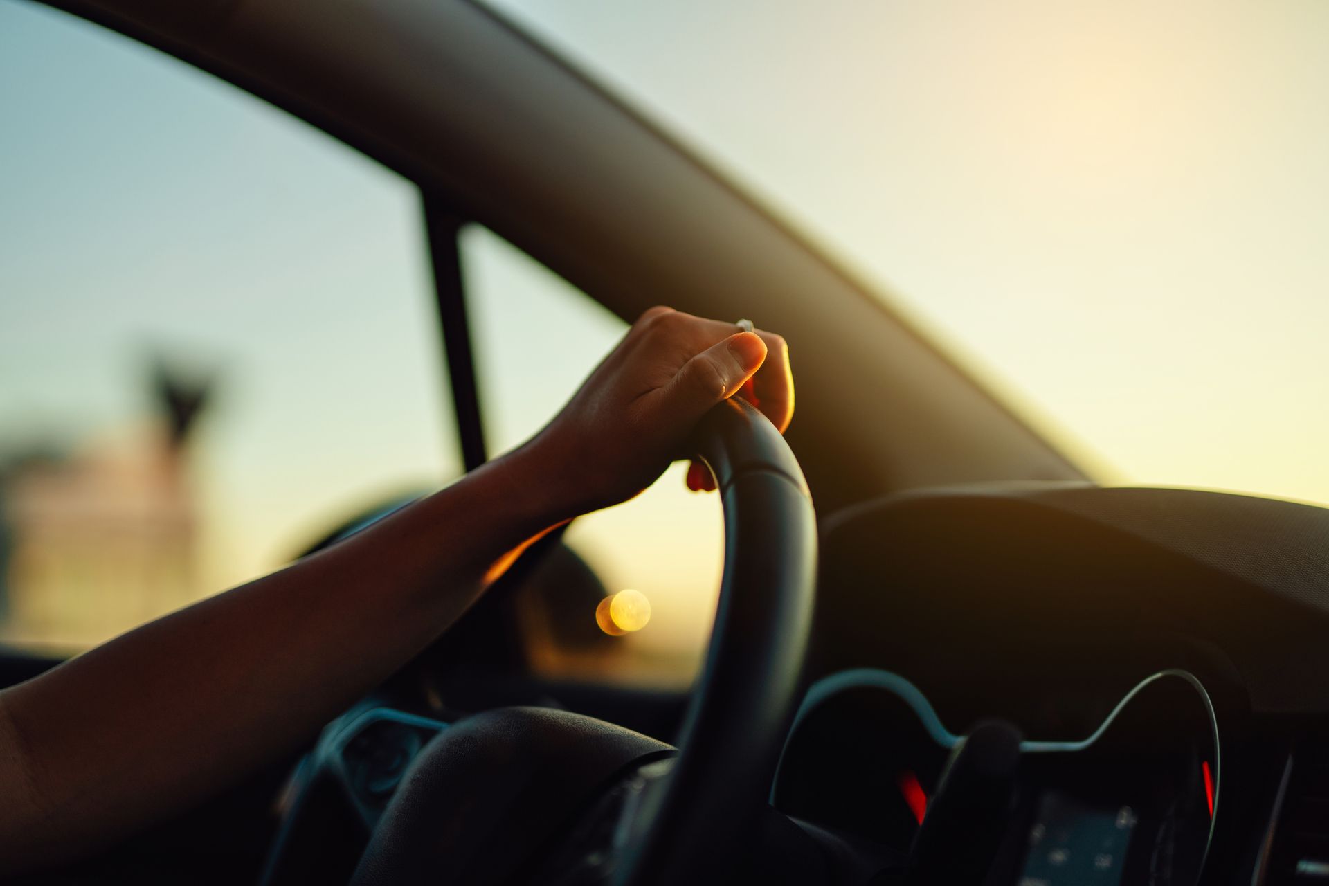 Hand gripping a steering wheel inside a car during sunset with warm light shining through.