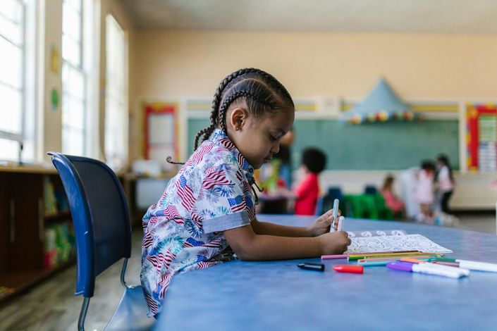 Child drawing at a table in a classroom, with markers and a chalkboard in the background