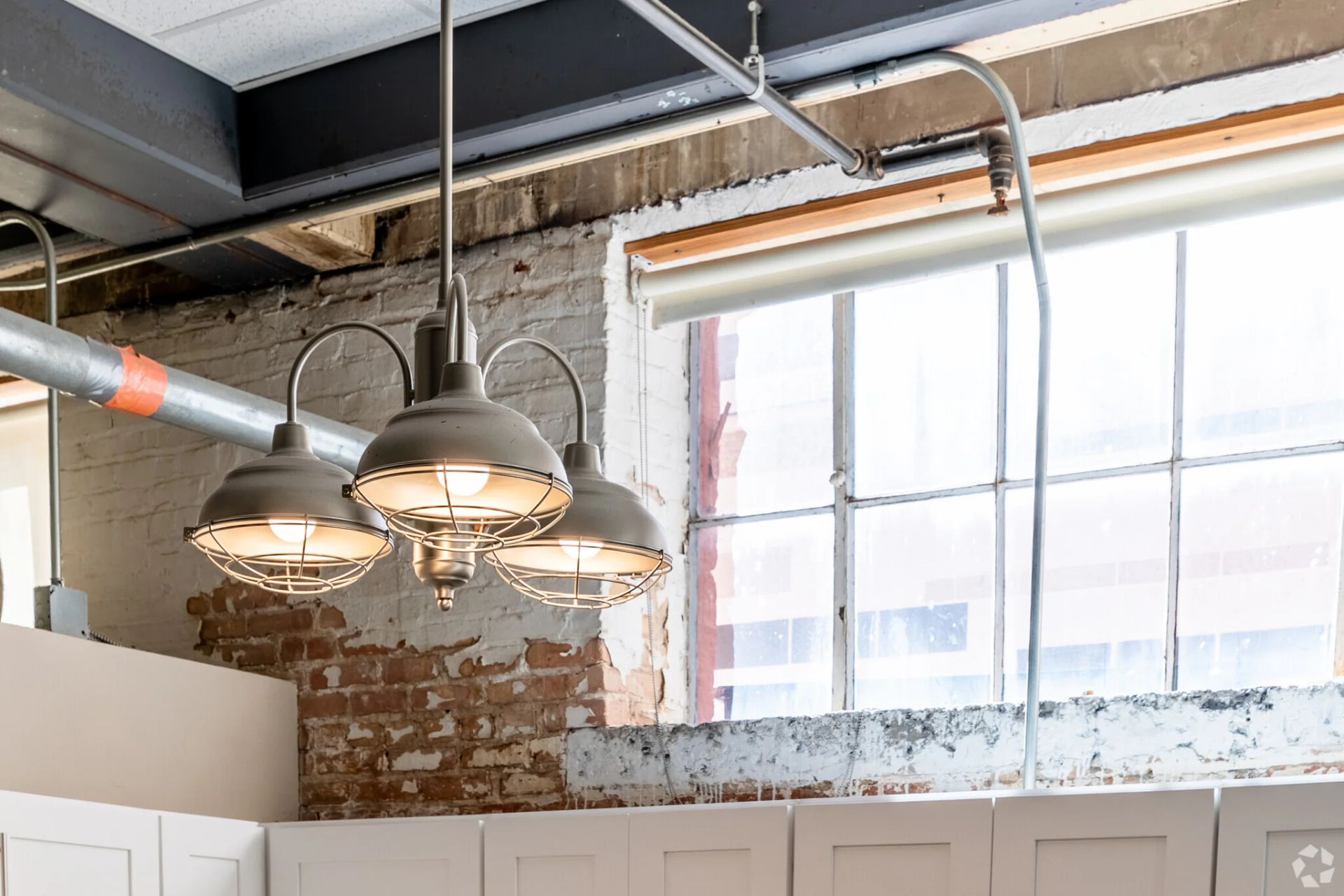 Industrial-style light fixture hanging in front of a brick wall and a large window.
