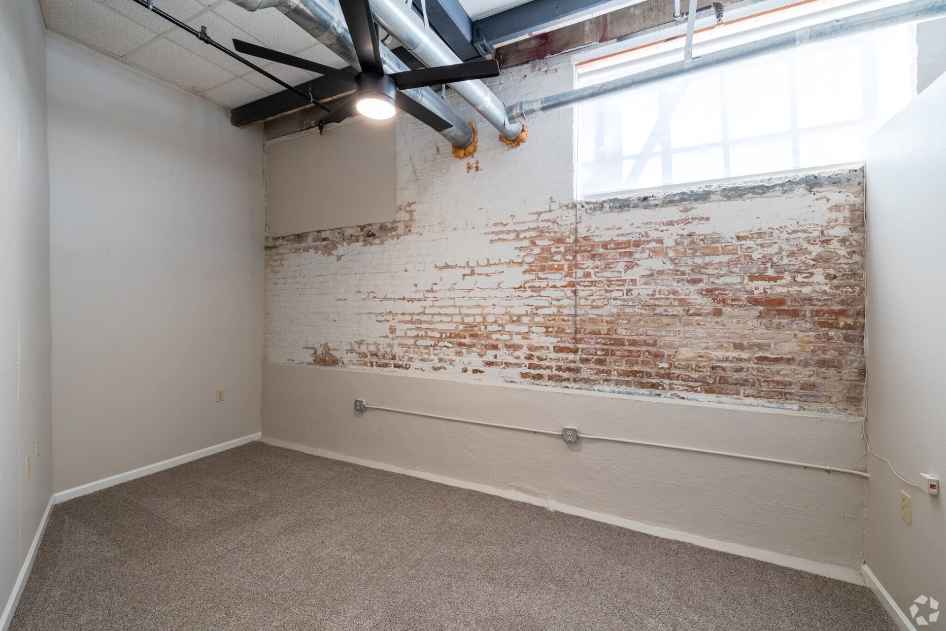 Empty room with exposed brick wall, carpeted floor, ceiling fan, and large window.