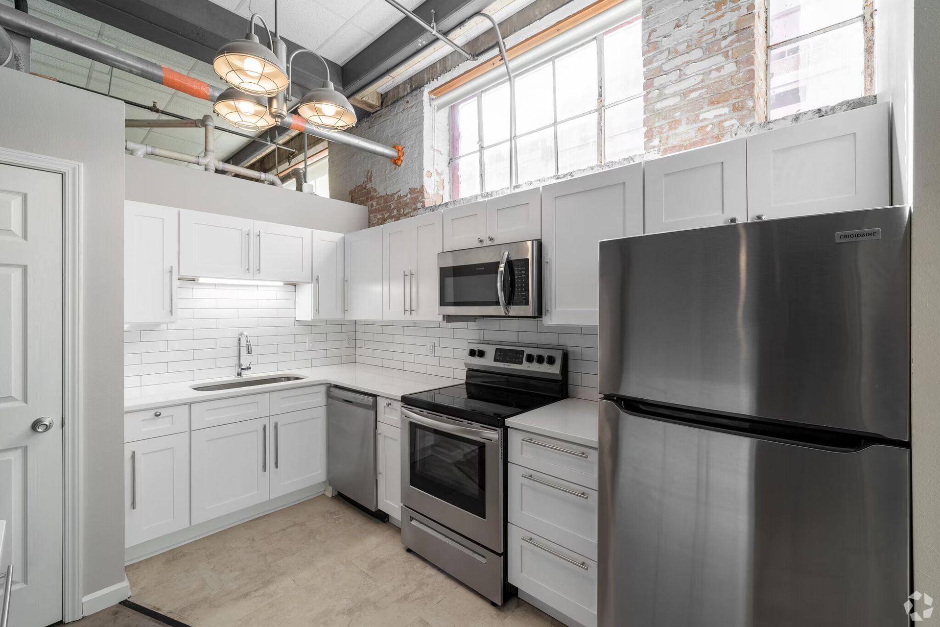 Modern white kitchen with stainless steel appliances, exposed brick, and high ceilings.