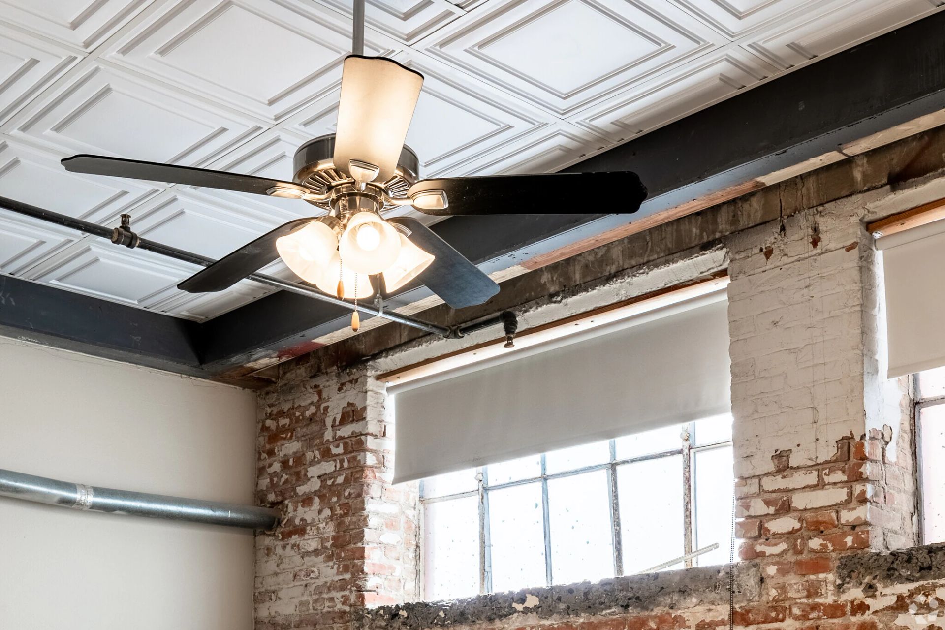 Ceiling fan with lights in a room with exposed brick and large windows.