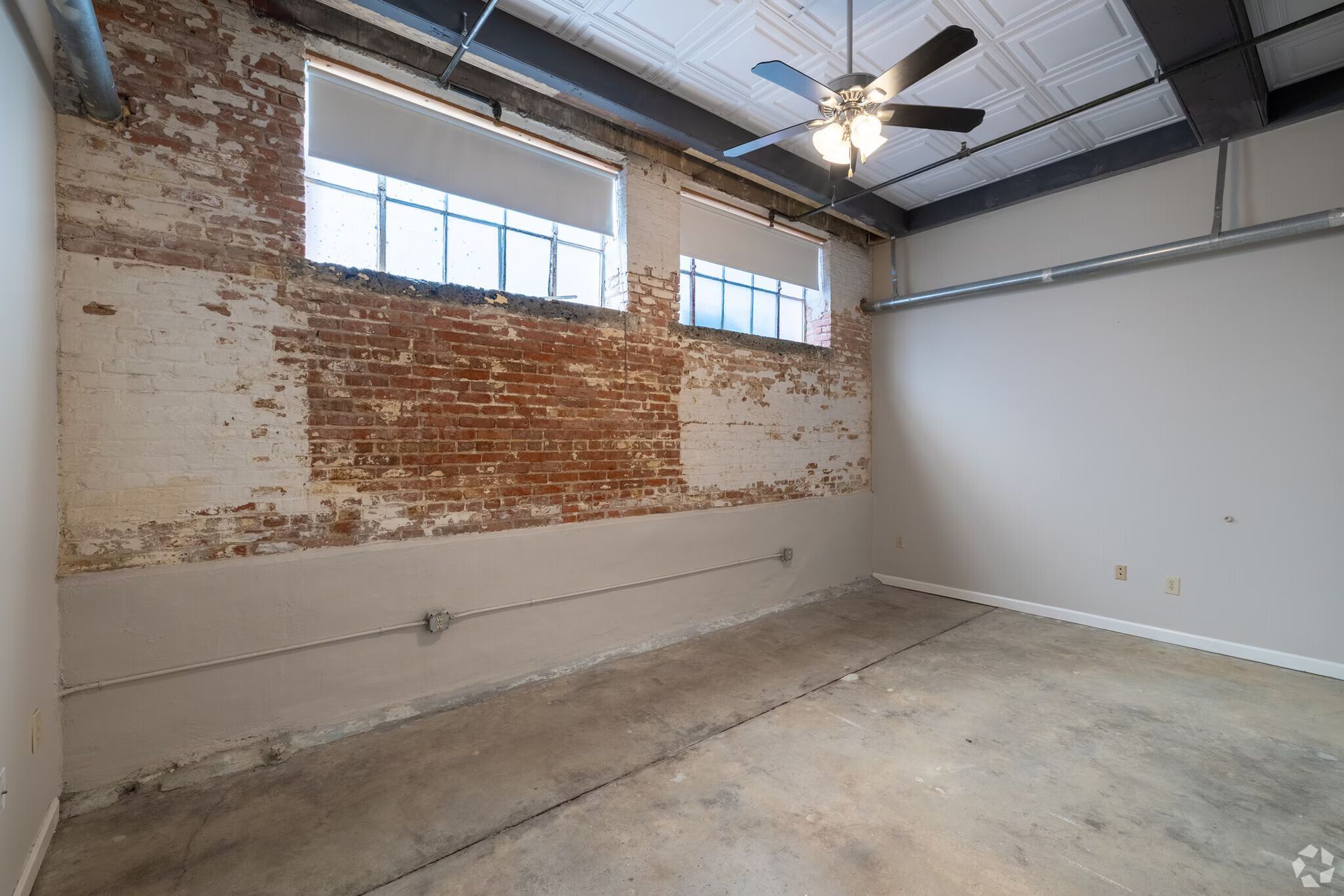 Interior view of an empty room with exposed brick wall, windows, concrete floor, and ceiling fan.