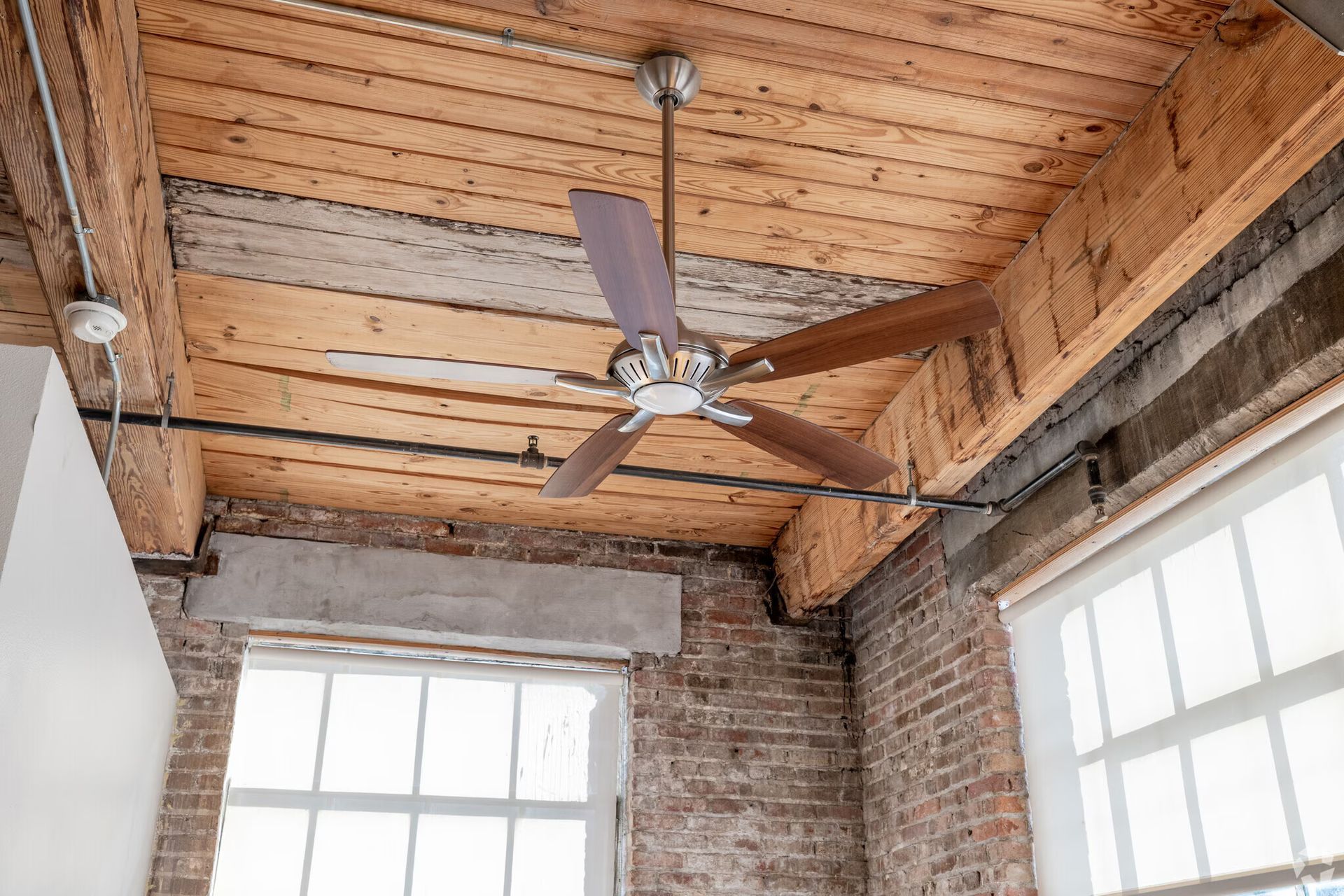 Ceiling fan in a room with exposed wooden beams, brick walls, and two windows.