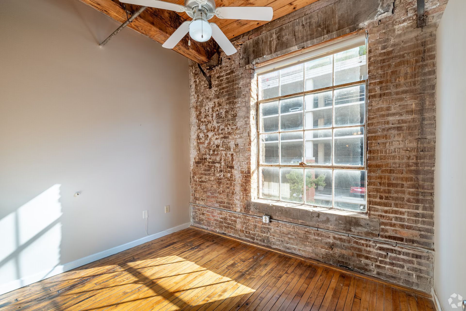 Empty room with exposed brick wall, window, wooden floor, and ceiling fan.