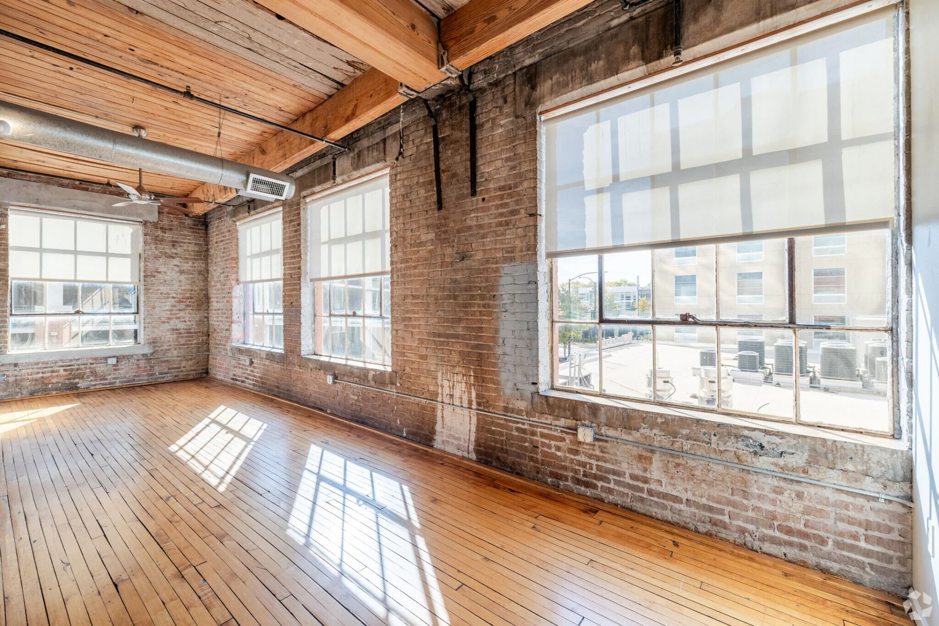 Loft interior with exposed brick walls, large windows, and wooden floors. Sunlight streams in.