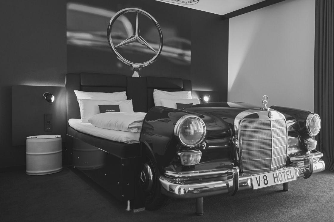 Ein orangefarbener Oldtimer wird in einem eleganten, modernen Ausstellungsraum mit dunklen Sitzen und einer Wandmalerei im Hintergrund präsentiert.