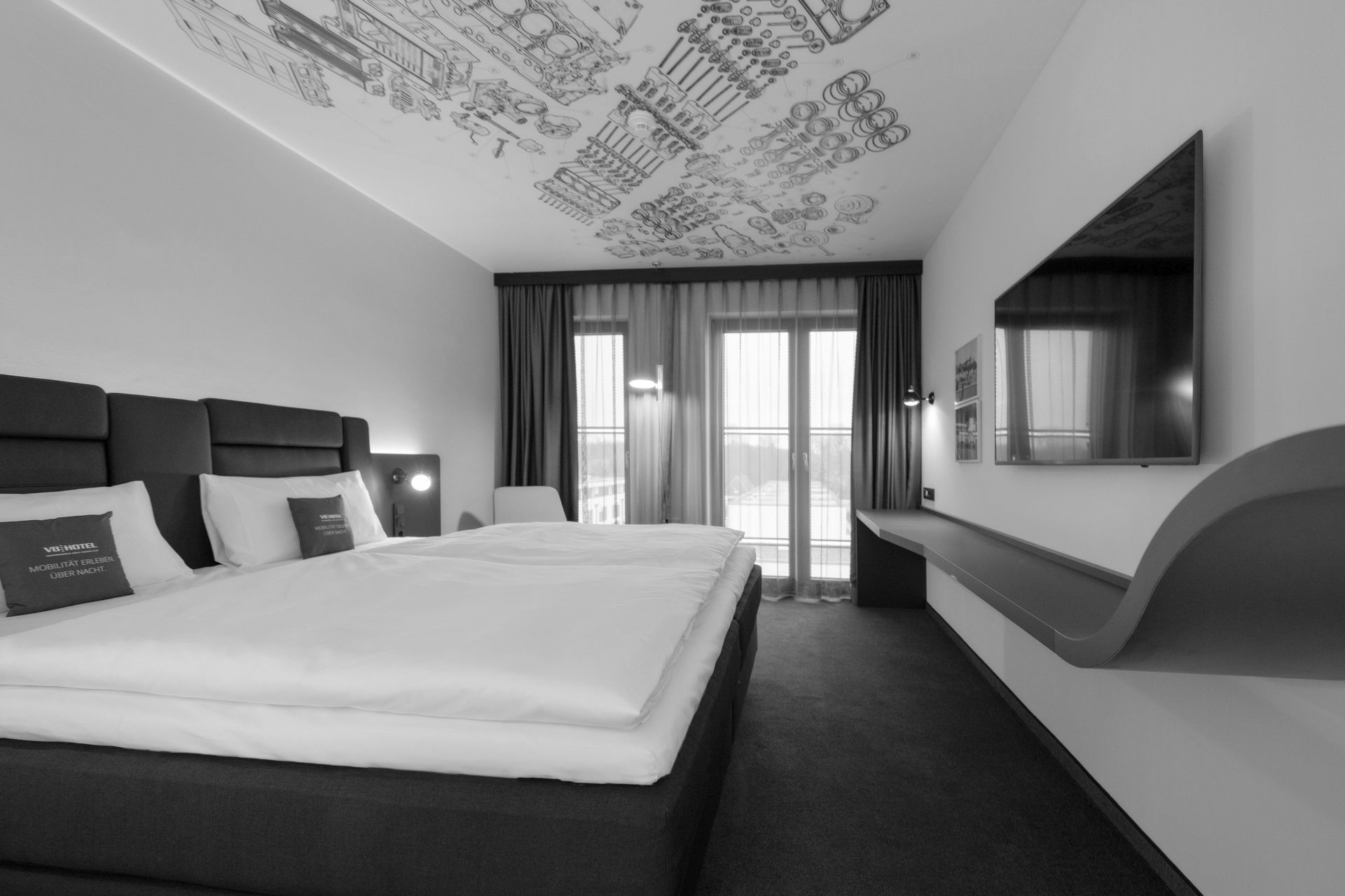 Modernes Hotelzimmer mit großem Bett, blauem Teppich, Fernseher und Balkontüren mit Stadtblick