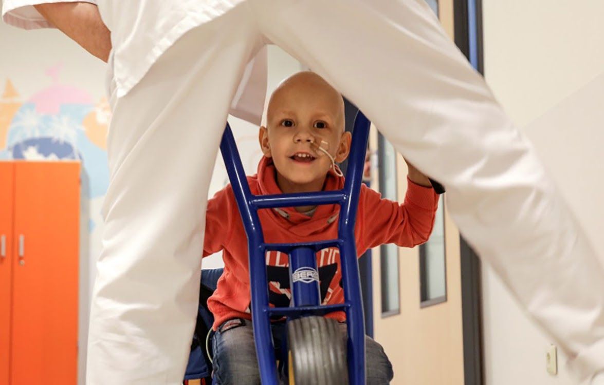 Een kind met een kaal hoofd, lachend, rijdend op een blauwe driewieler, ondersteund door een persoon in een wit uniform in een felgekleurde kamer.