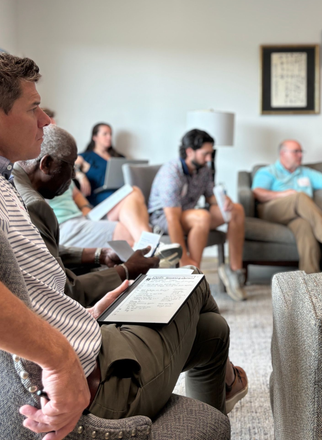 Group therapy session in a rustic room. People sitting in a circle, one person gestures with a paper.