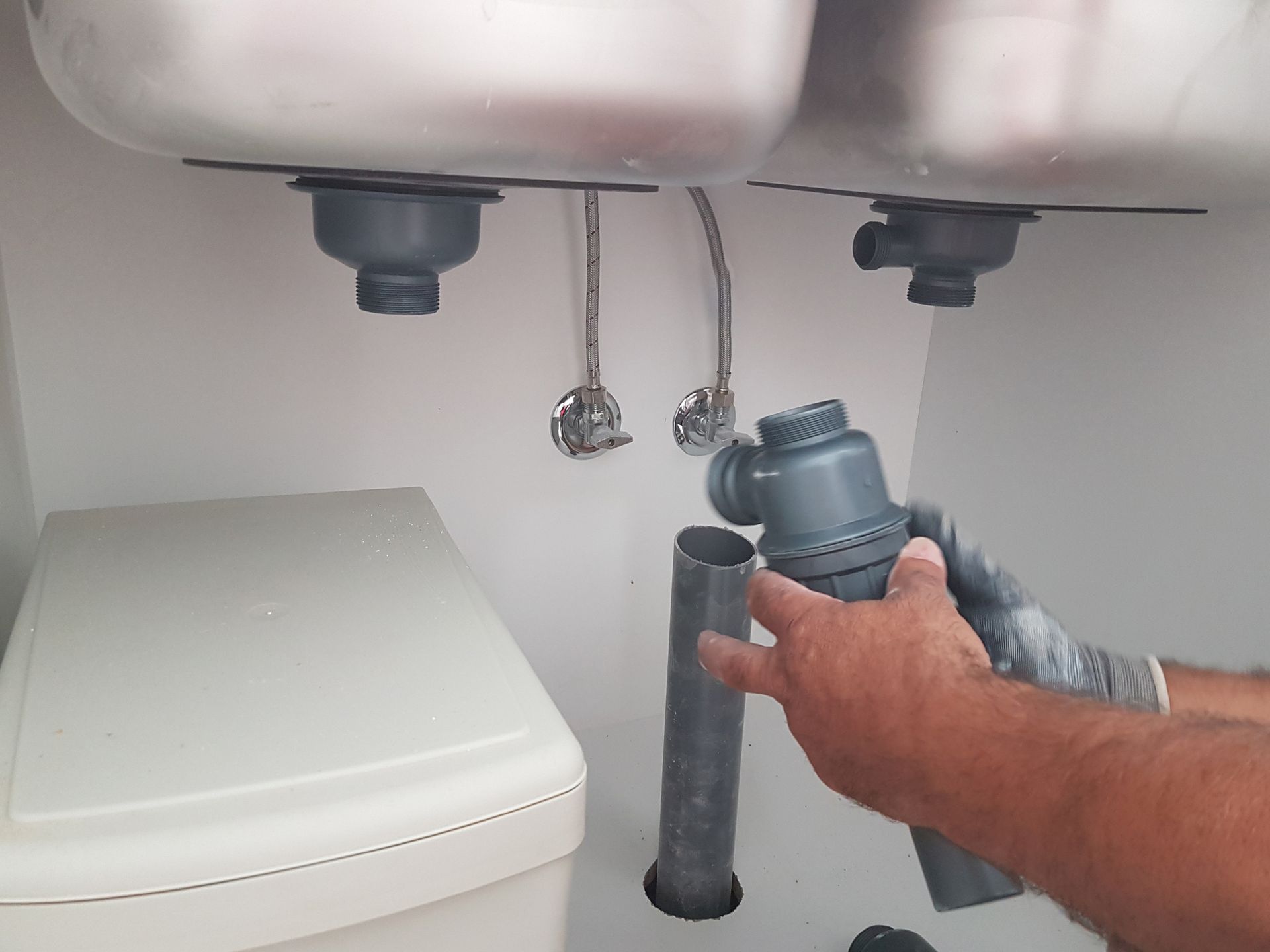Person holding a sink trap, with drain pipes and sinks visible Person holding a sink trap, with drain pipes and sinks visible