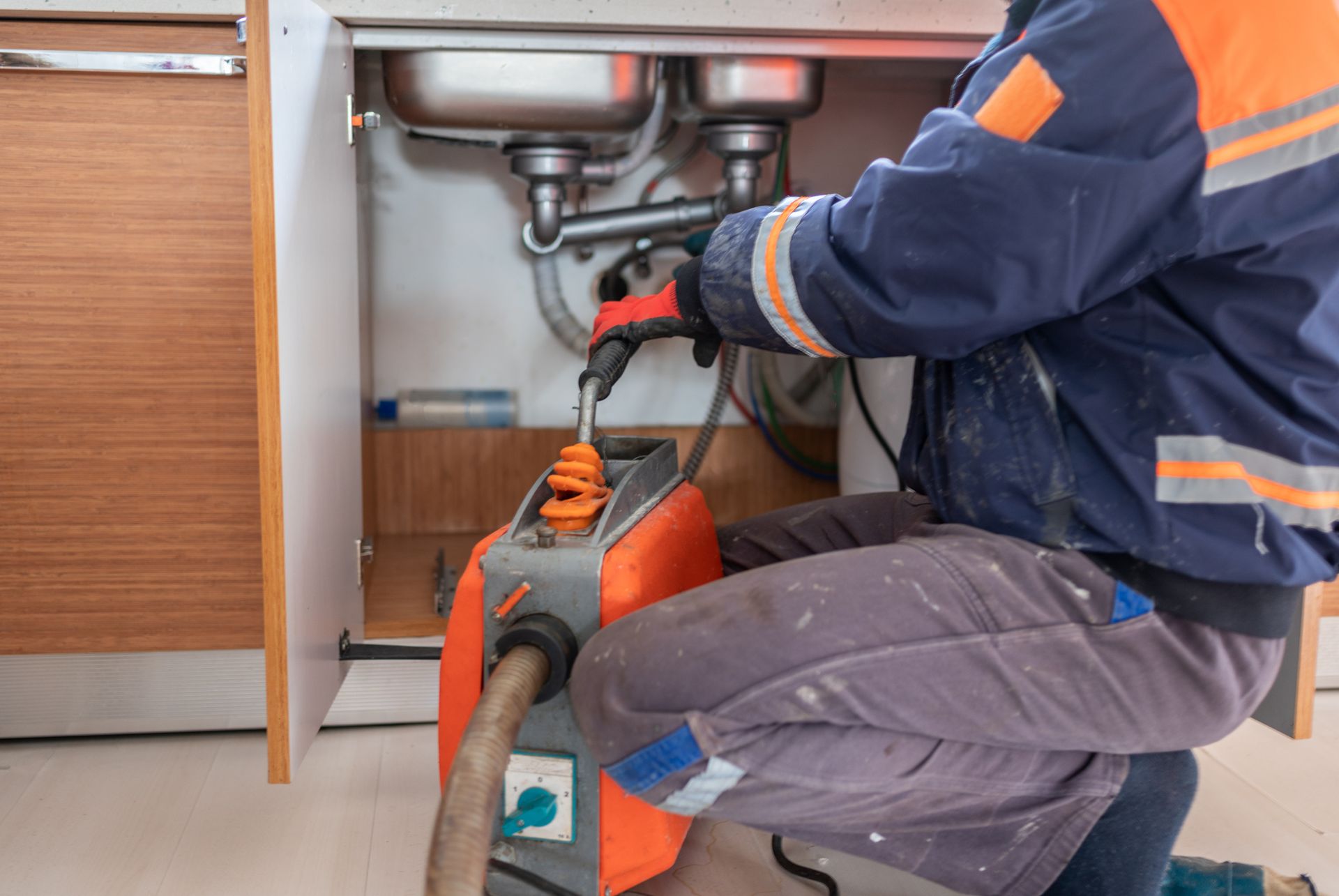 A person using a drain cleaning machine under a kitchen sink A person using a drain cleaning machine under a kitchen sink