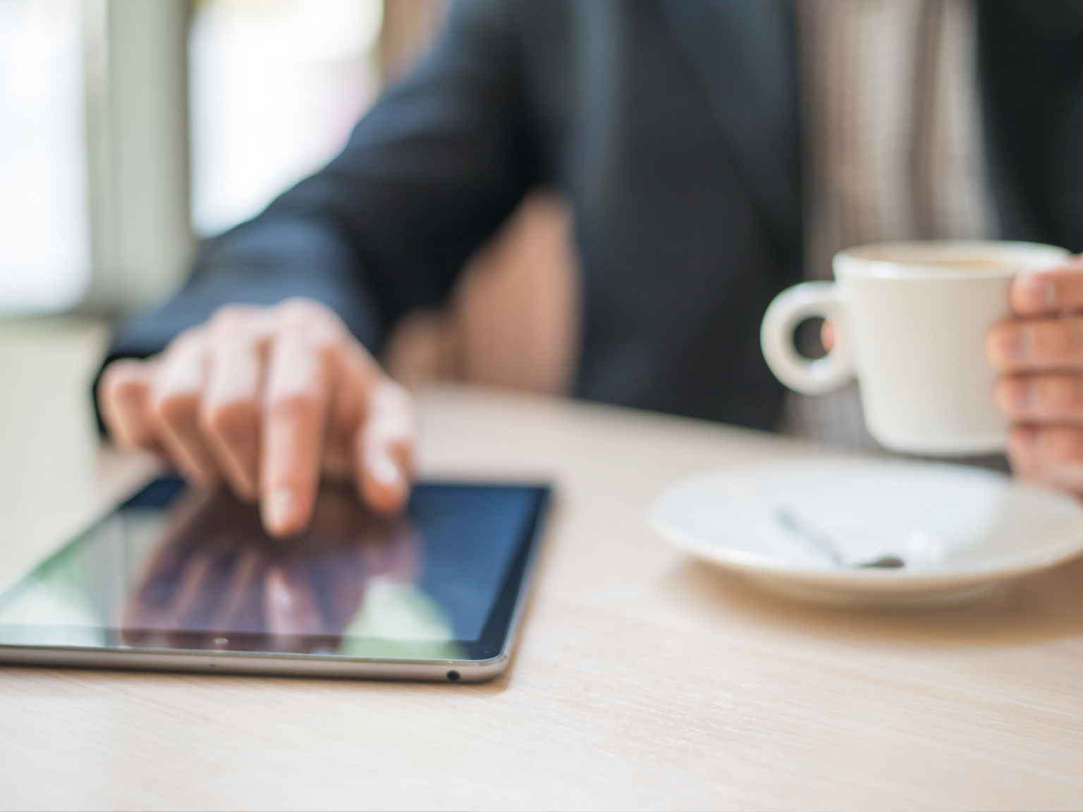 A man is sitting at a table using a tablet computer while holding a cup of coffee.
