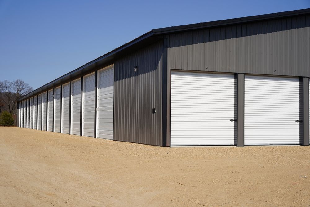 Long row of gray storage units with white roll-up doors on a gravel lot under a clear sky