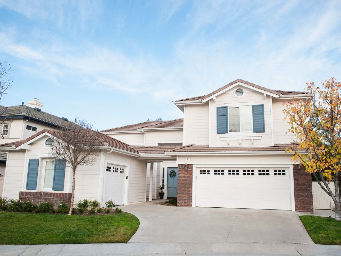 Suburban two-story beige house with white garage, blue shutters, and a small front lawn