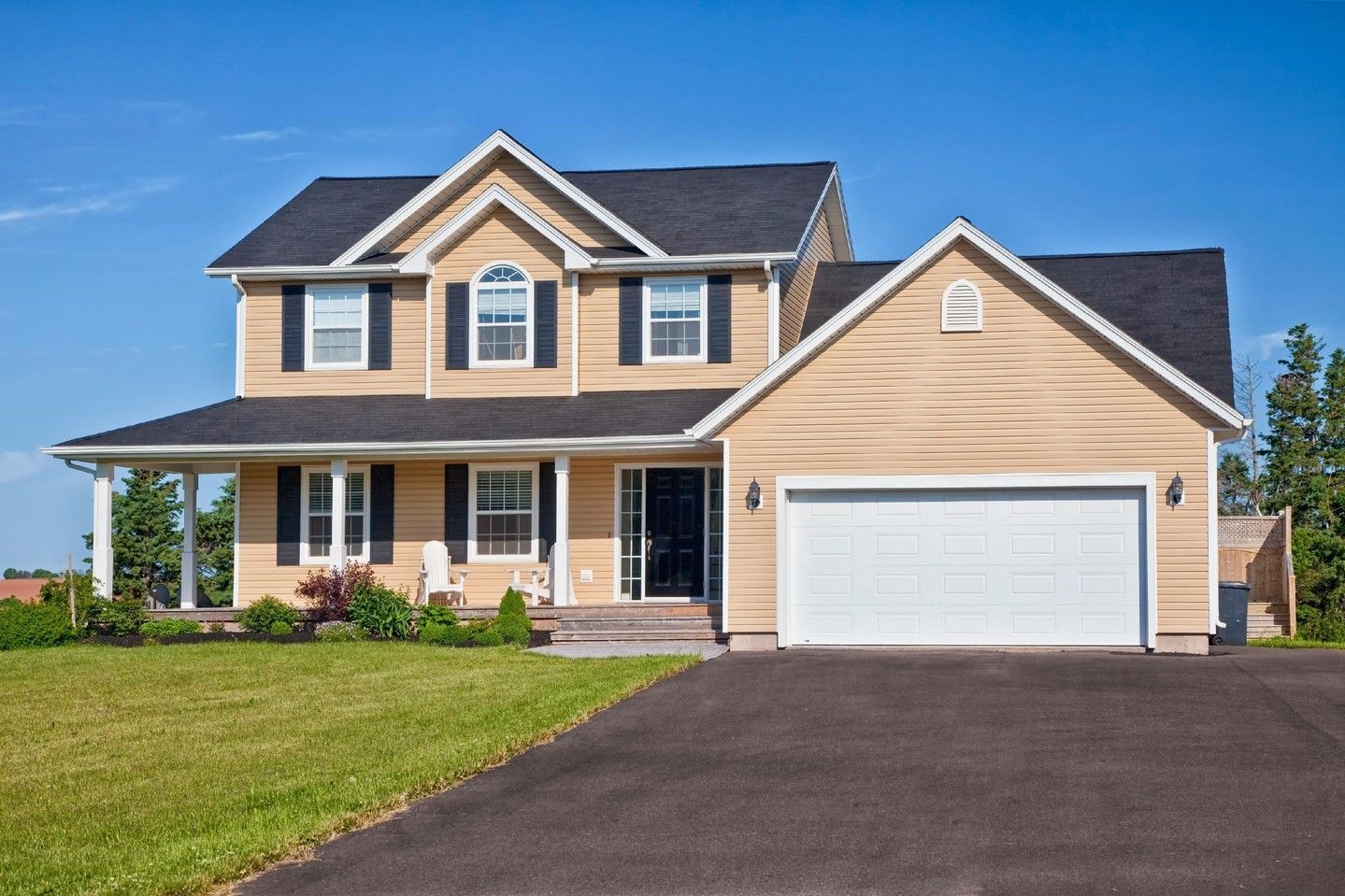 Suburban two-story beige house with white garage and front porch, viewed from the driveway under a blue sky
