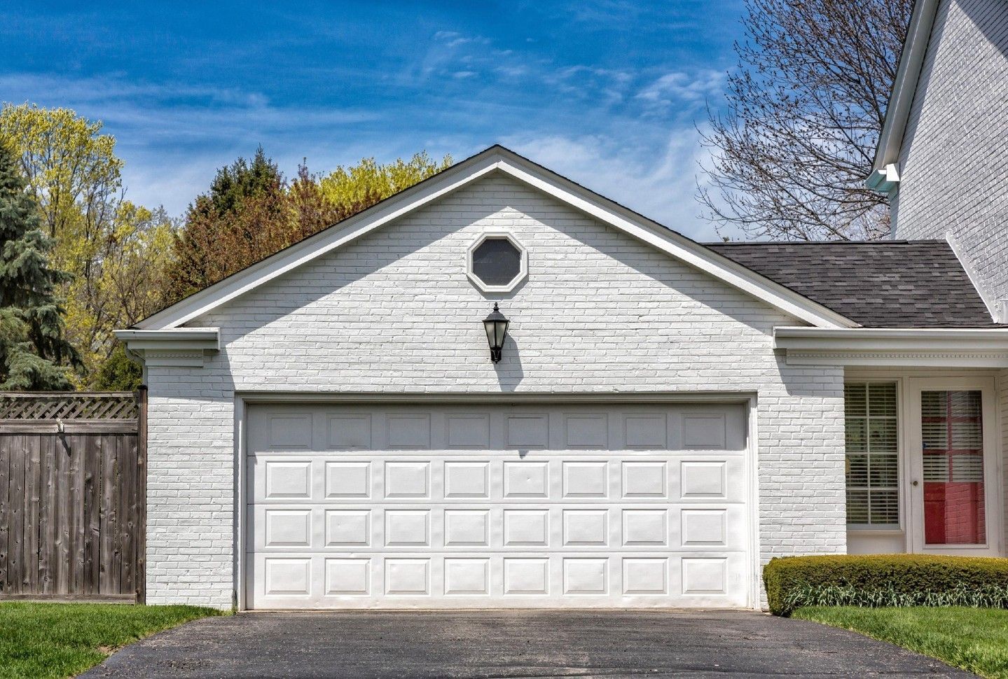 White detached garage with double door and small octagonal window, beside a house and driveway under a blue sky