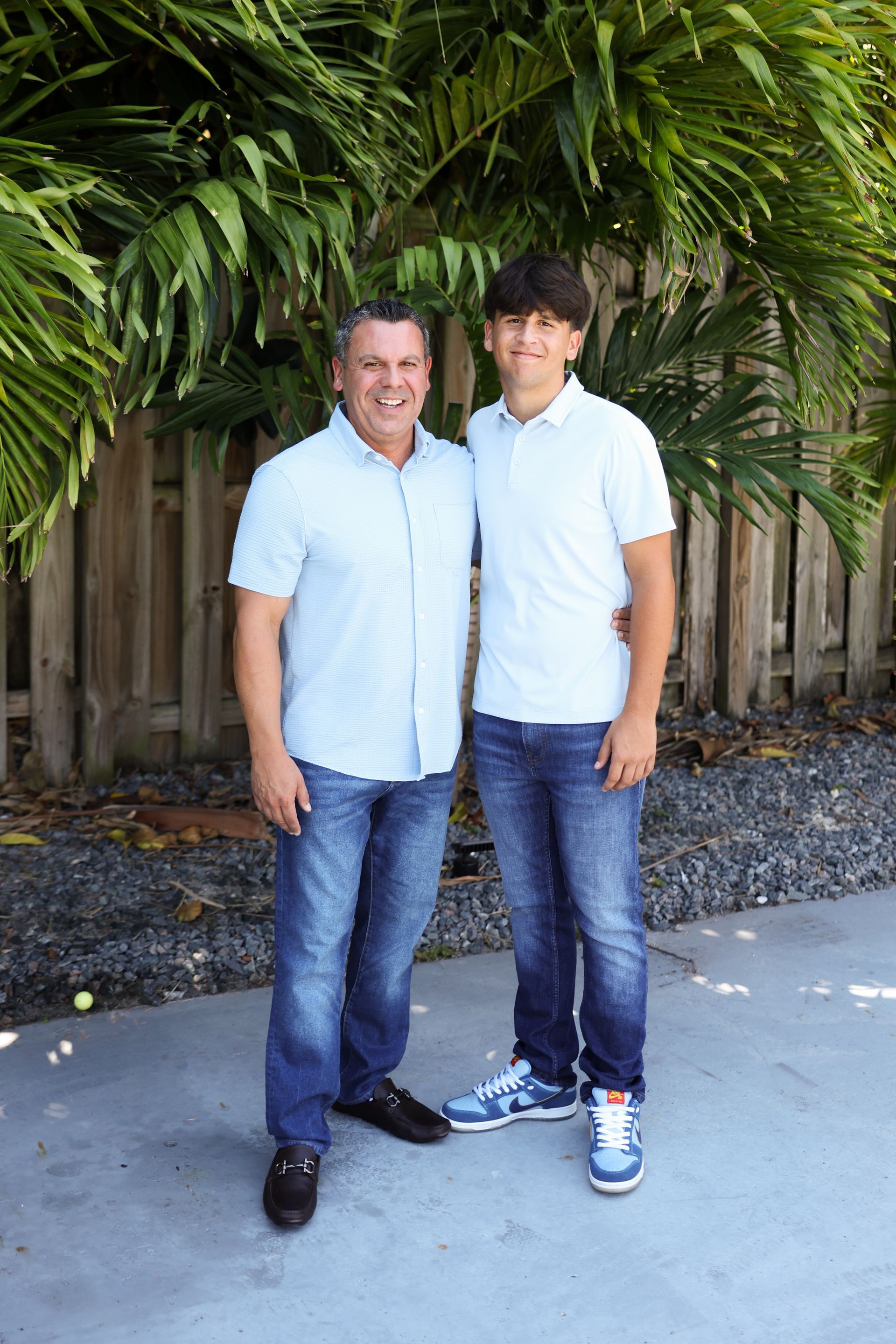A man and a boy are standing next to each other in front of a fence.
