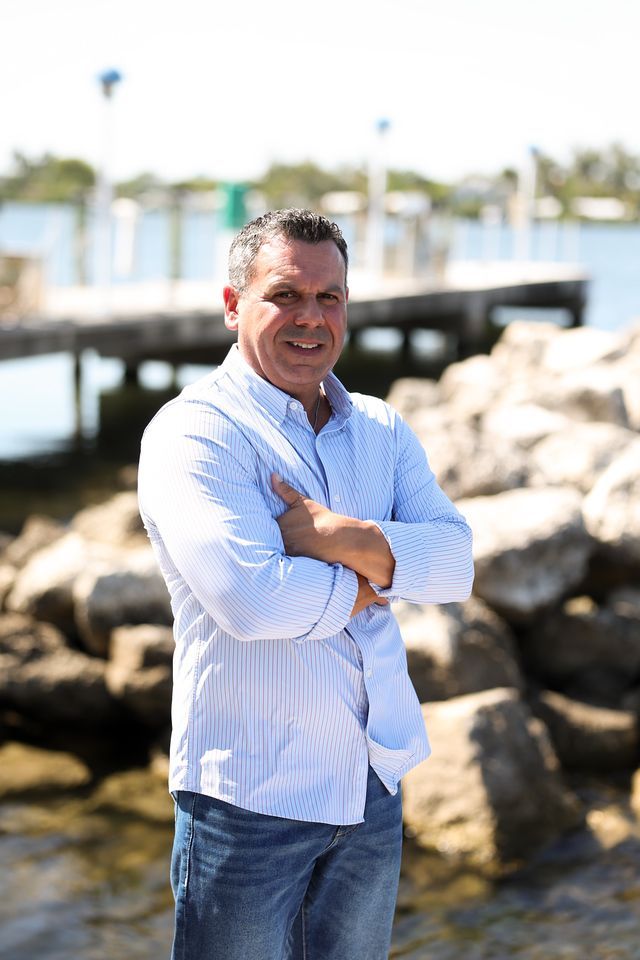 A man in a white shirt and blue jeans is standing in front of a body of water with his arms crossed.