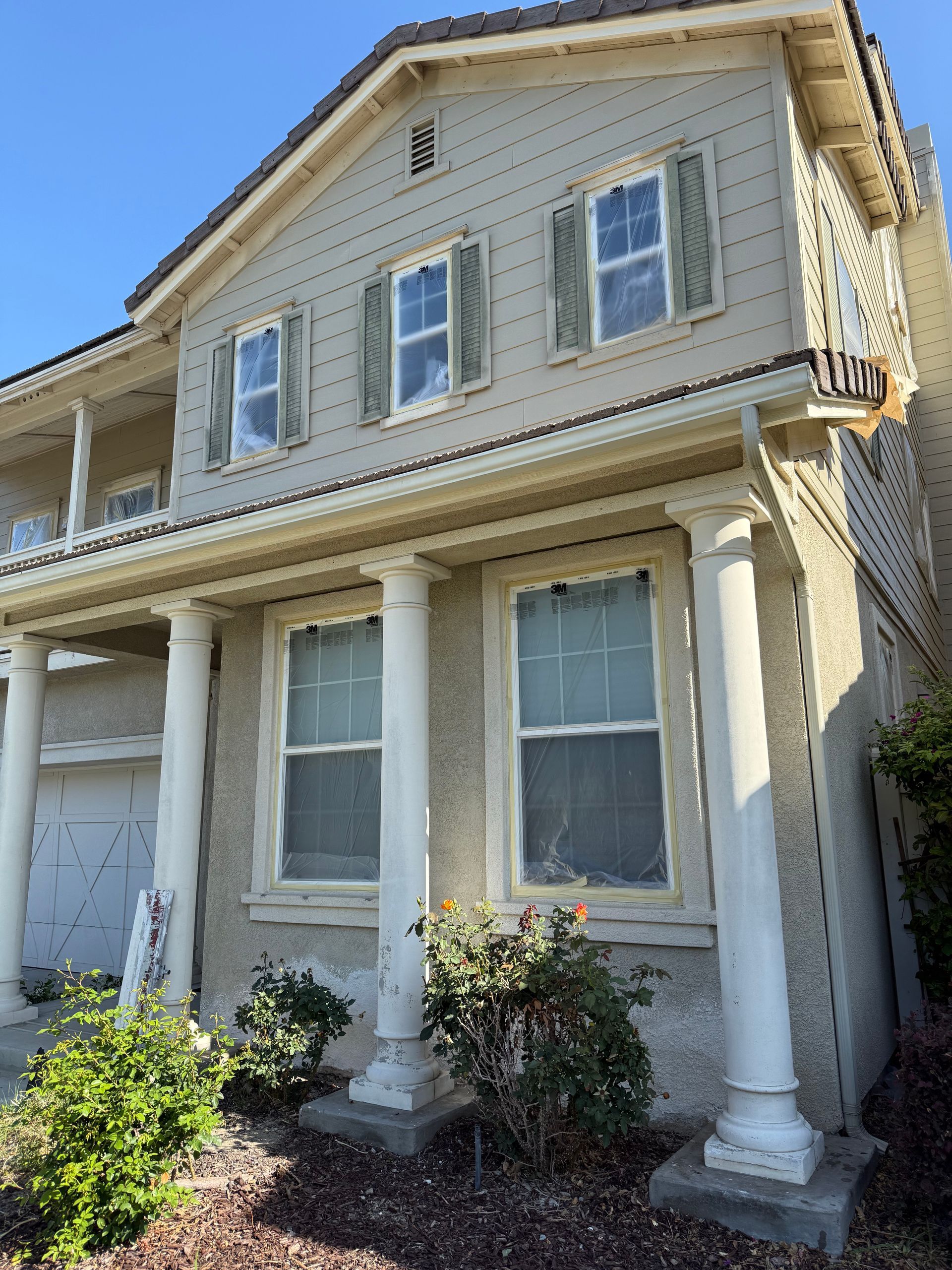 Freshly painted Two-story beige house with white pillars and green shutters. Bushes and a garage door are visible on the ground floor.
