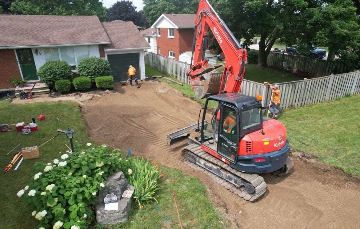 A red excavator sits on a partially cleared dirt driveway next to a residential house with a lawn and fence.