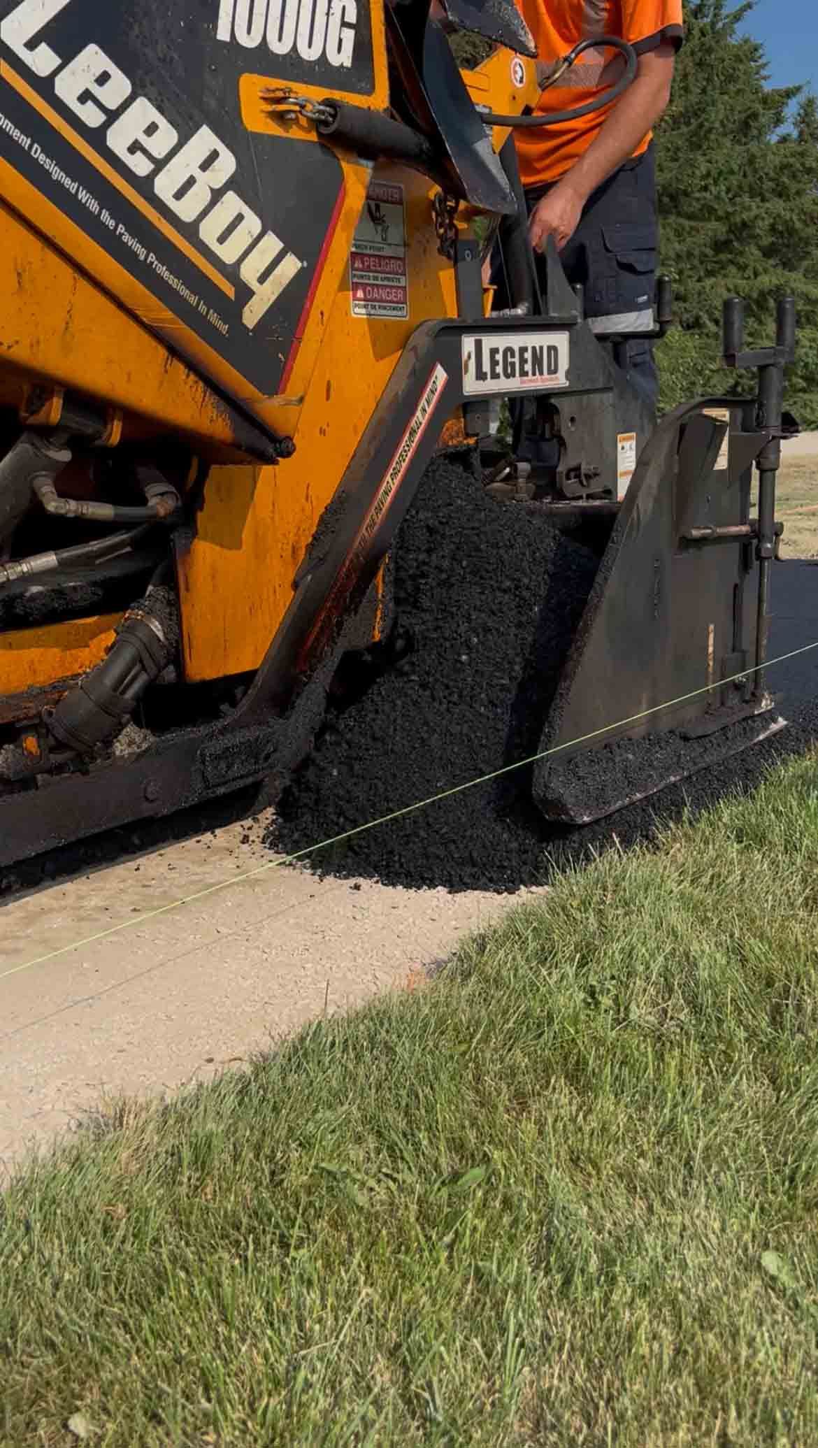 A yellow LeeBoy paving machine deposits black asphalt onto a gravel road surface.