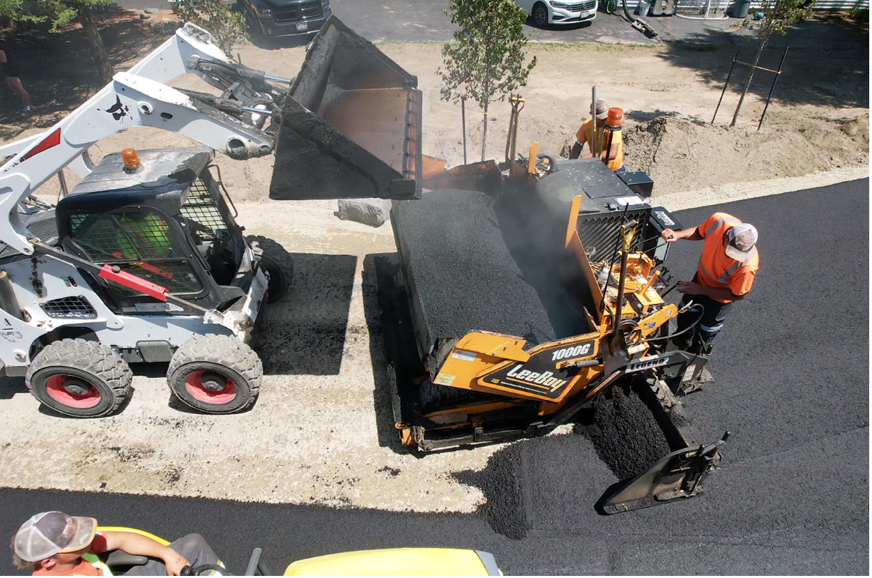 A skid-steer loader and a paving machine work together to lay fresh asphalt on a construction site.