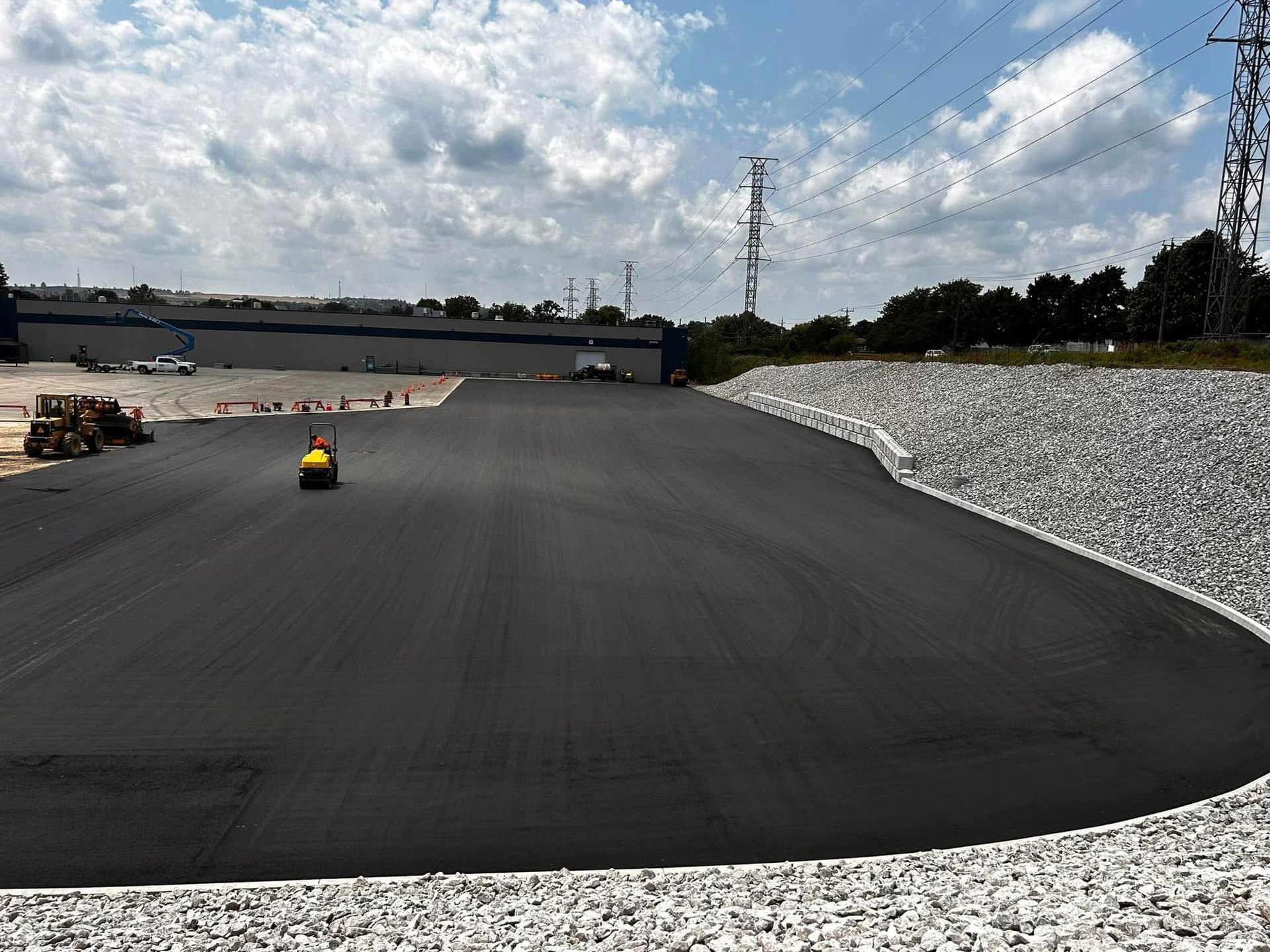 A steamroller levels fresh black asphalt in a large parking lot next to a stone embankment under a sunny, cloudy sky.