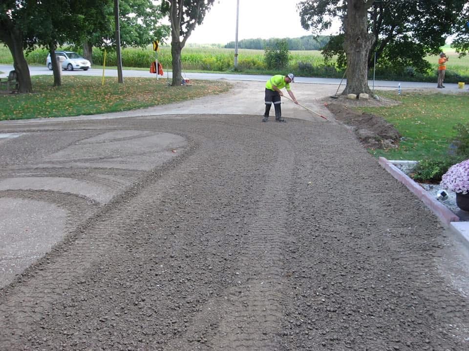A construction worker in a high-visibility yellow shirt rakes gravel over a residential driveway.
