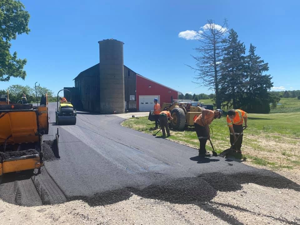 Workers in safety gear pave a new asphalt driveway leading to a red barn and silo on a sunny day.