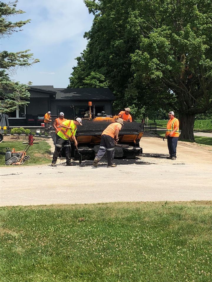 A crew in high-visibility orange vests works to pave a driveway with machinery in front of a dark house.