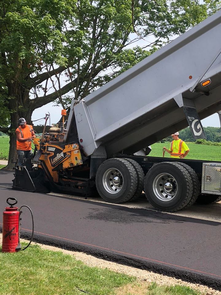 A dump truck feeds asphalt into a road paver on a sunny day, with two workers nearby on a grassy roadside.