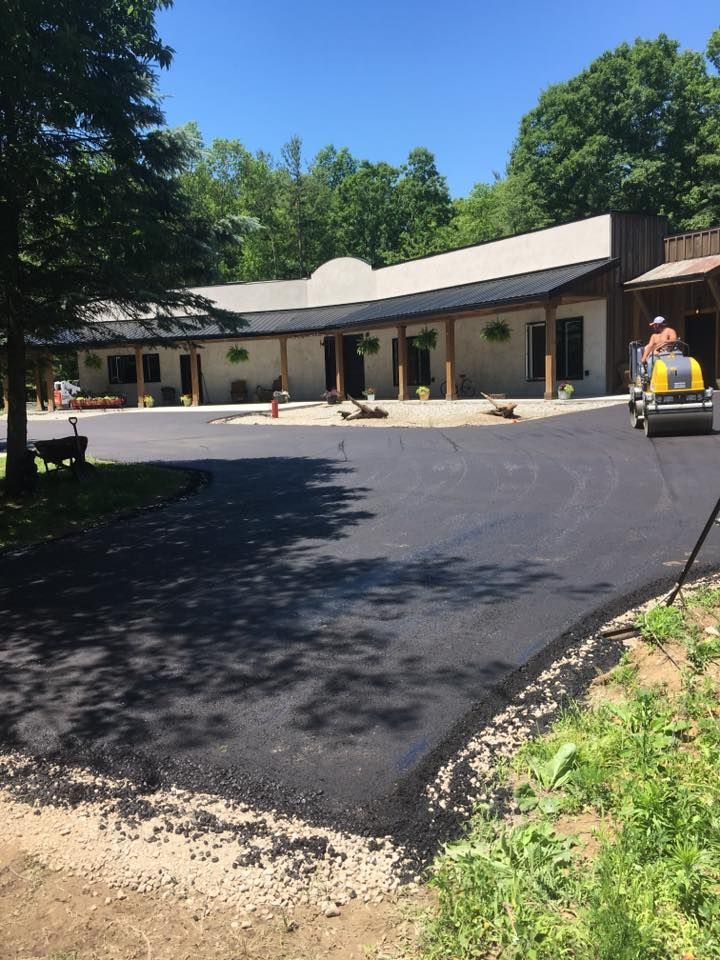 A construction worker operates a steamroller on a newly paved black asphalt parking lot in front of a tan commercial building.