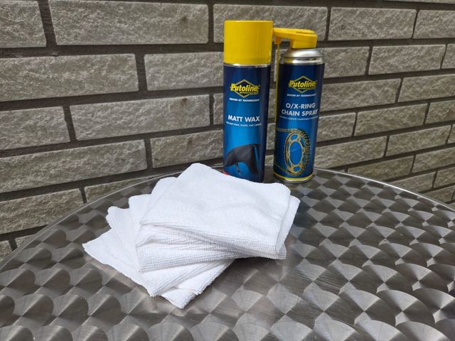 Twee spuitbussen, schoonmaakdoeken op een metalen tafel, bakstenen muur op de achtergrond.