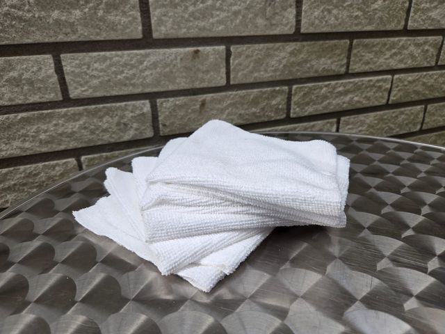 Een stapel witte, opgevouwen washandjes op een zilverkleurige tafel met patroon, tegen een bakstenen muur op de achtergrond.