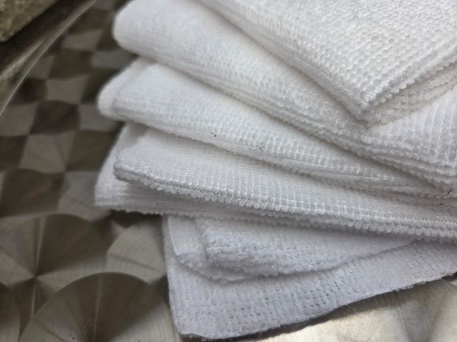 Een stapel witte, opgevouwen washandjes op een zilverkleurige tafel met patroon, tegen een bakstenen muur op de achtergrond.