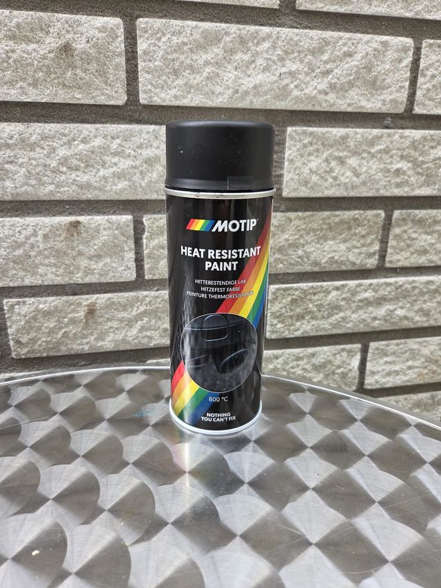Zwarte MOTIP hittebestendige verf spuitbus op een reflecterend metalen oppervlak, voor een bakstenen muur.