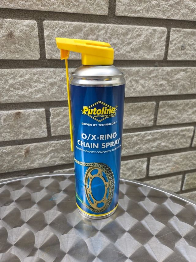 Een bus Putoline O/X-ring kettingspray met een gele spuitmond staat op een tafel met een patroon voor een bakstenen muur.