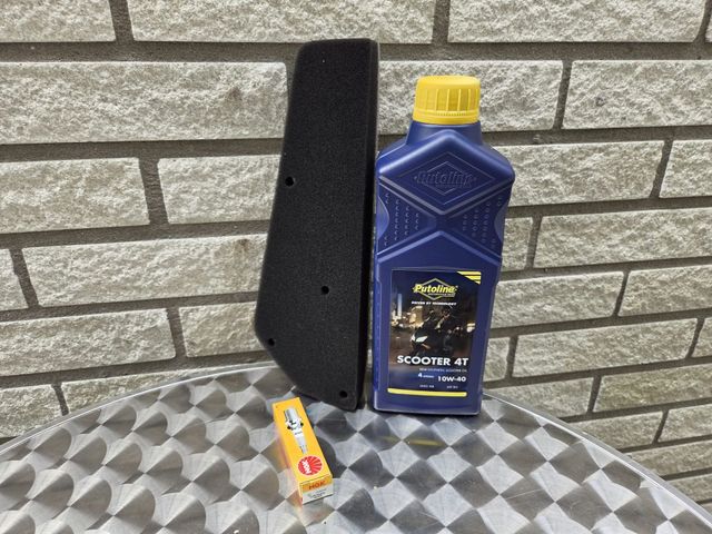 Een zwart luchtfilter, een fles olie en een bougie liggen op een geruite tafel voor een bakstenen muur.