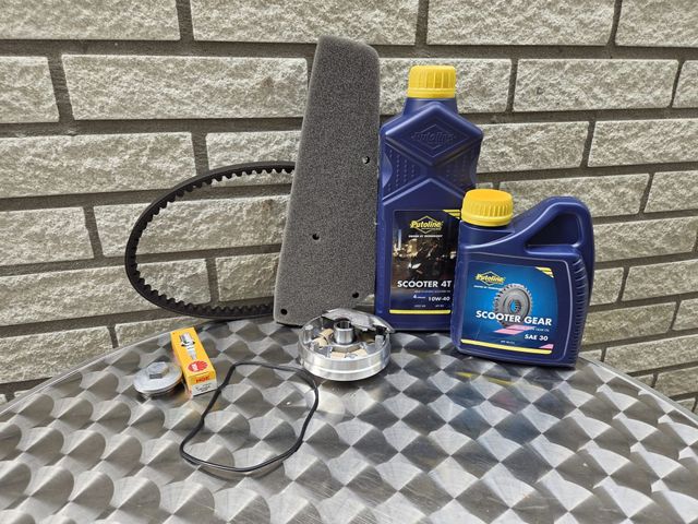 Motoronderdelen: riem, luchtfilter, olie, koppelingsset, op een geruite tafel tegen een bakstenen muur.