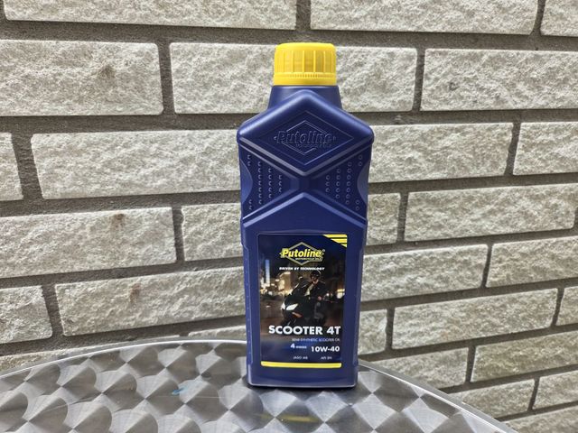 Blauwe fles scooterolie met gele dop, op een metalen ondergrond tegen een bakstenen muur.