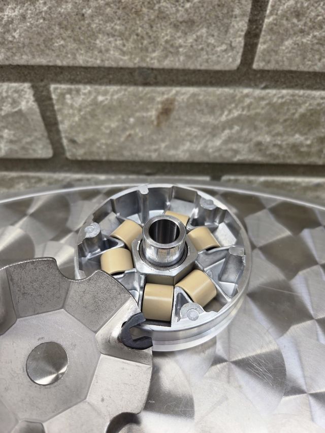 Een scootervariator met beige rollen en een metalen tandwiel op een glanzend oppervlak, tegen een bakstenen muur.