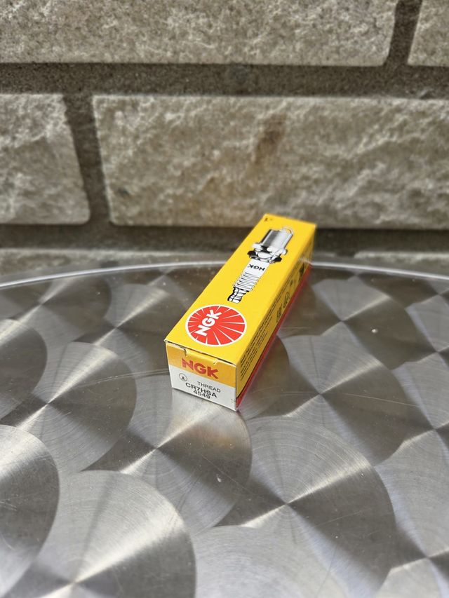 Een gele NGK-bougiedoos op een zilverkleurige, gestructureerde ondergrond, voor een grijze bakstenen muur.