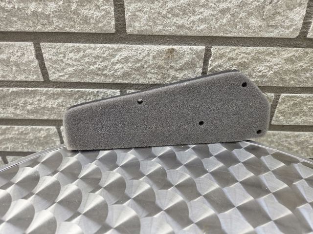 Grijs schuimrubberen luchtfilter op een metalen oppervlak met patroon, tegen een bakstenen muur op de achtergrond.