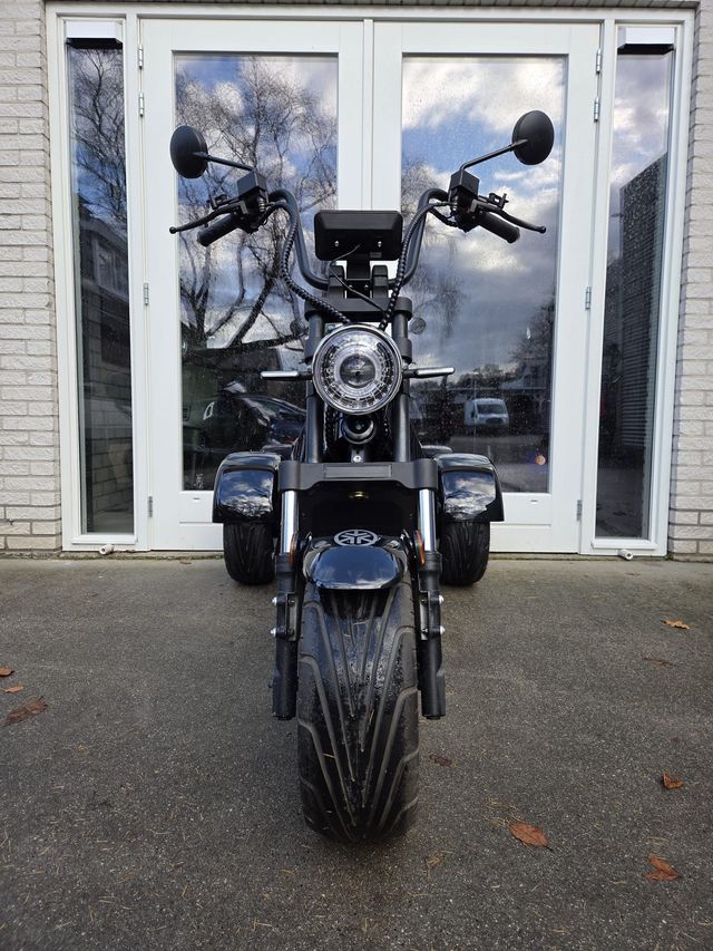 Een zwarte driewielige scooter staat geparkeerd voor glazen deuren; hij heeft een zadel, een stuur en een laadruimte.