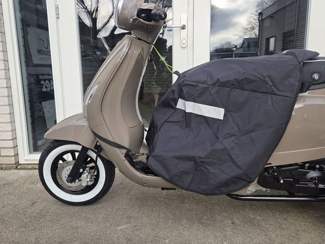 Persoon op een beige scooter met beenbeschermers. Witte banden, reflecterende strip op de beschermer, geparkeerd voor een gebouw.