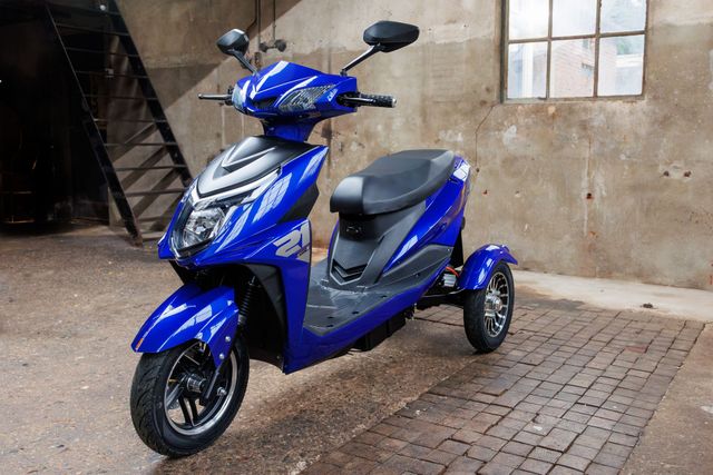 Een blauwe elektrische driewieler staat binnen geparkeerd, vlakbij een betonnen muur en een metalen trap.