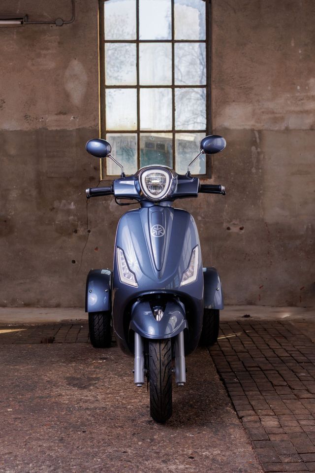 Een blauwe driewielige scooter staat geparkeerd in een verweerd bakstenen gebouw, naast een raam.
