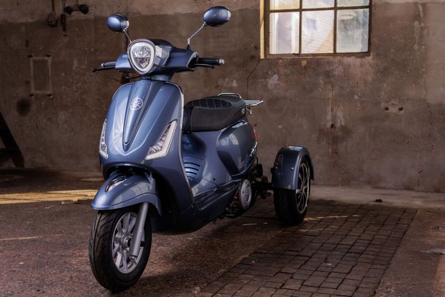 Een blauwe driewielige scooter staat geparkeerd in een verweerd bakstenen gebouw, naast een raam.