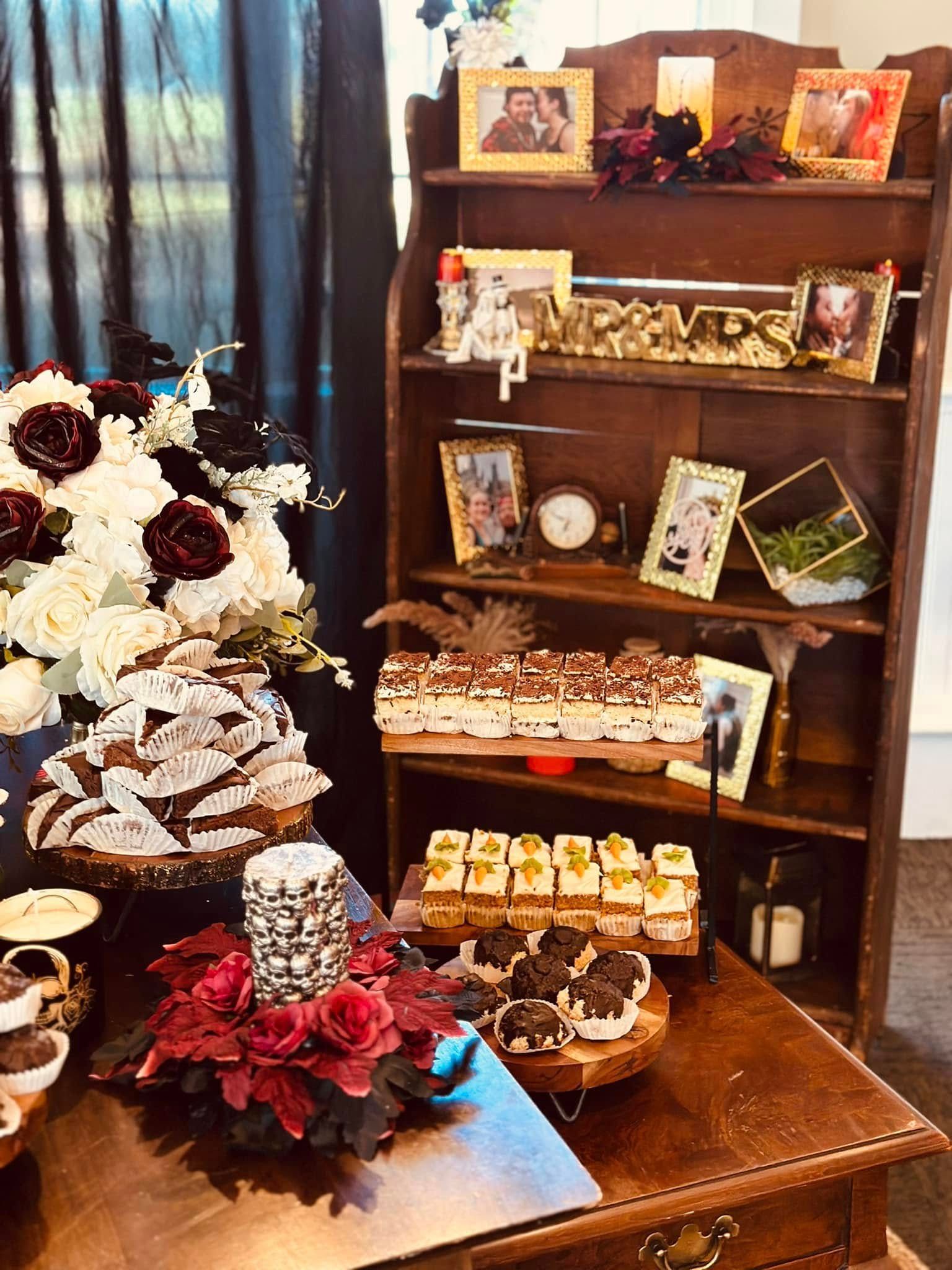 A table with a variety of desserts and flowers on it in front of a bookshelf.